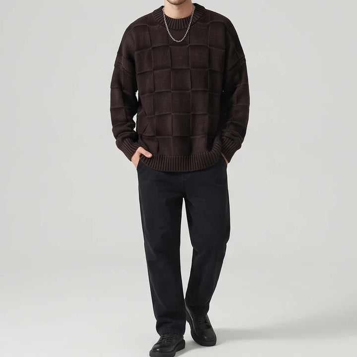 Man wearing an oversized brown knit sweater with a textured checkerboard pattern, black pants, and a silver chain necklace.