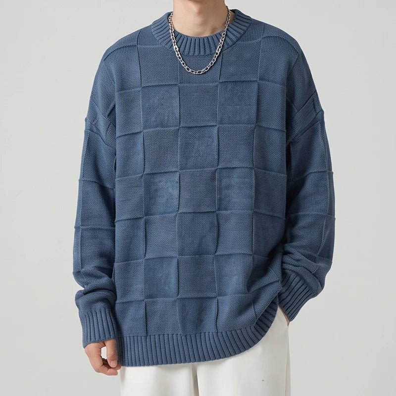 Man wearing an oversized indigo blue checker knit sweater with a silver chain, paired with white pants.