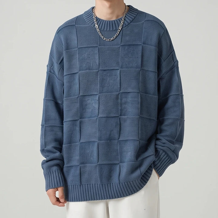 Man wearing an oversized indigo blue checker knit sweater with a silver chain, paired with white pants.