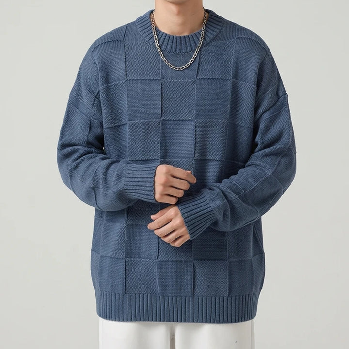 Man wearing an oversized indigo blue knit sweater with a textured checkerboard pattern and a ribbed hem, posed against a plain background.