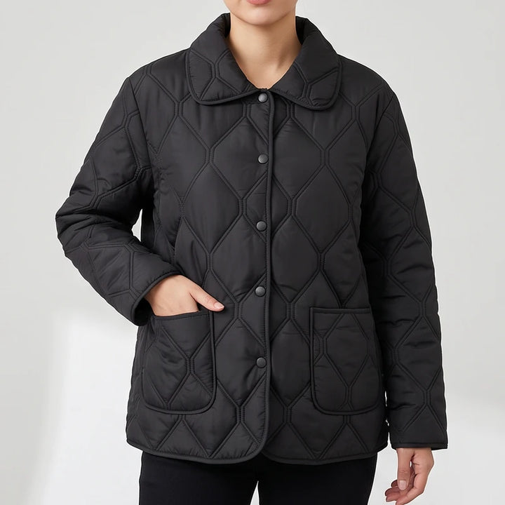 Woman wearing a black quilted jacket with a pointed collar, round buttons, and two front patch pockets, one hand in a pocket.