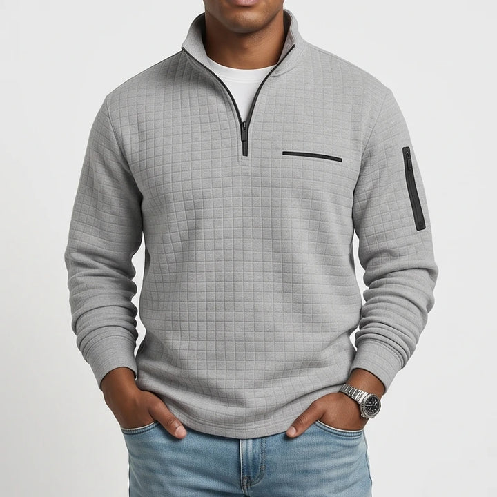Man wearing a light grey quilted quarter-zip pullover with black zippers and subtle chest detail, hands in pockets.