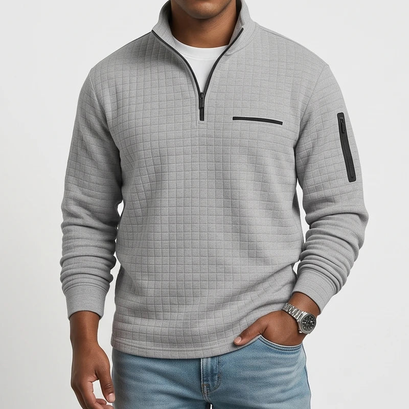 Man wearing a gray quilted pullover with a black quarter-zip, chest pocket, sleeve zipper, and light blue jeans.