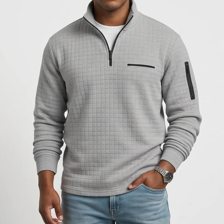 Man wearing a gray quilted pullover with a black quarter-zip, chest pocket, sleeve zipper, and light blue jeans.