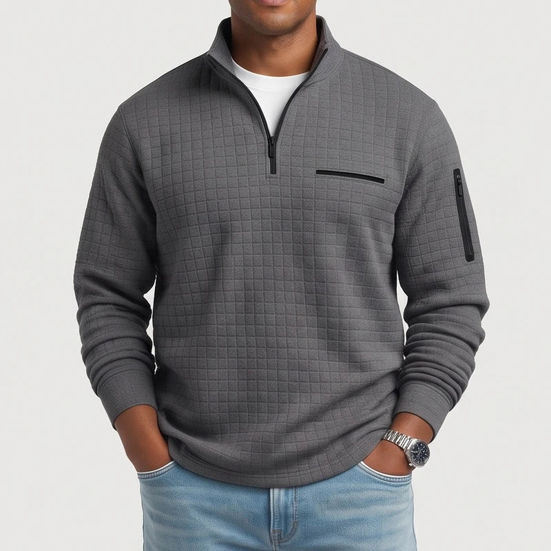 Man wearing a dark gray quilted quarter-zip pullover with a chest pocket and sleeve pocket, hands in light wash jeans.