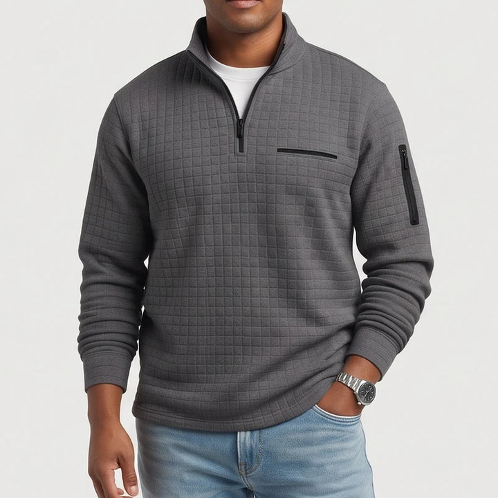Man wearing a dark gray quilted quarter-zip pullover with black zippers, a chest pocket, and an arm pocket.
