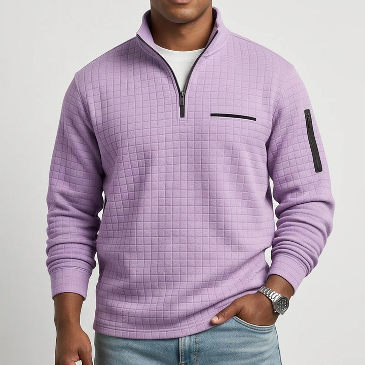 Man wearing a lavender quilted pullover with a black quarter-zip, chest pocket, and sleeve zipper, hand in jeans pocket.