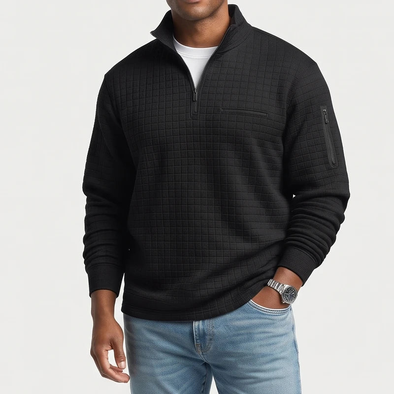 Man wearing a black quilted quarter-zip pullover with chest pocket, arm zipper pocket, and white undershirt, with hand in light wash jeans.
