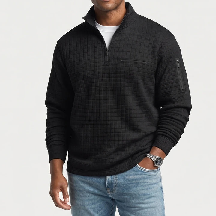 Man wearing a black quilted quarter-zip pullover with chest pocket, arm zipper pocket, and white undershirt, with hand in light wash jeans.