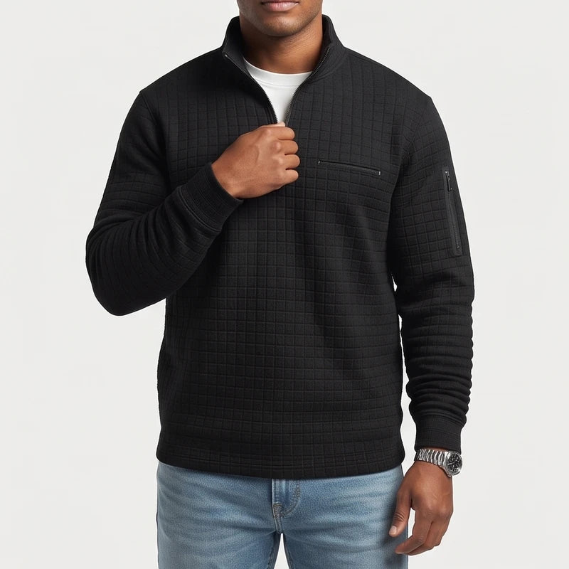 Man wearing a black quilted quarter-zip pullover with chest and sleeve zipper pockets, light wash jeans, and a silver watch.