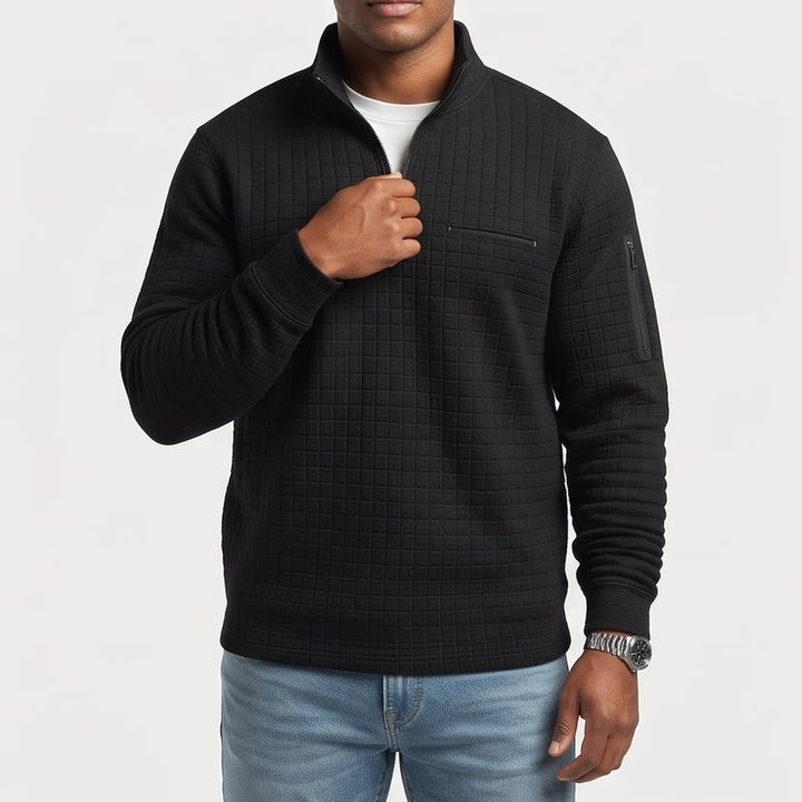 Man wearing a black quilted quarter-zip pullover with chest and sleeve zipper pockets, light wash jeans, and a silver watch.