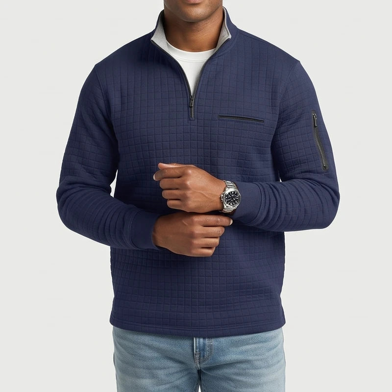Man wearing a navy blue quilted quarter-zip pullover with chest and sleeve zipper pockets, light denim jeans, and a watch.