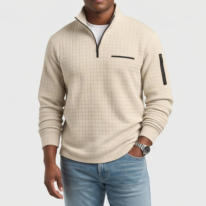 Man wearing a beige quilted quarter-zip pullover with black zippers, light blue jeans, and a silver watch.