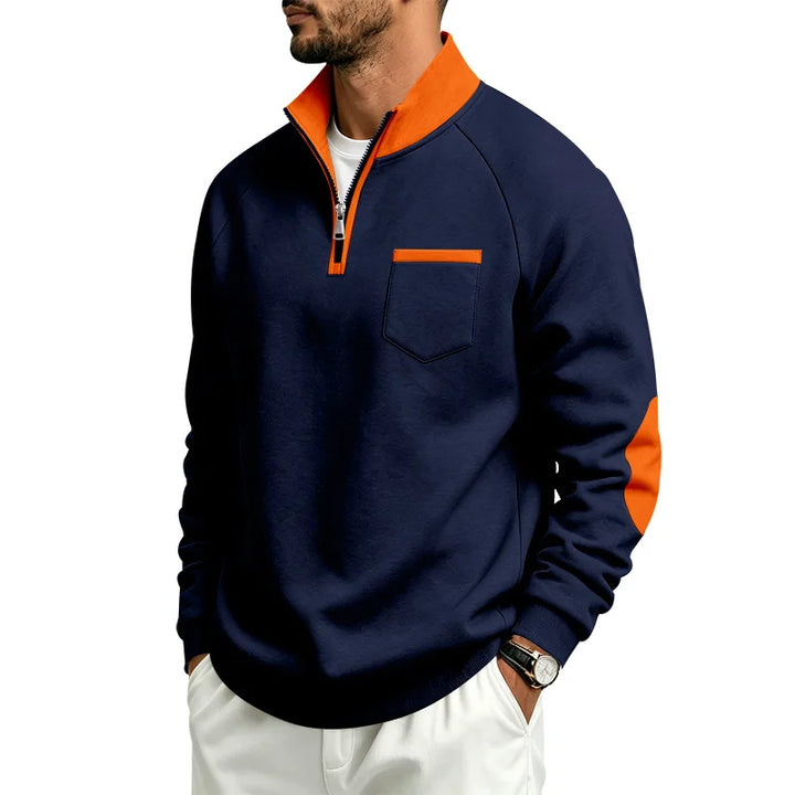 Man wearing a navy blue quarter-zip pullover with an orange collar, pocket trim, and elbow patches, posing with hands in pockets.