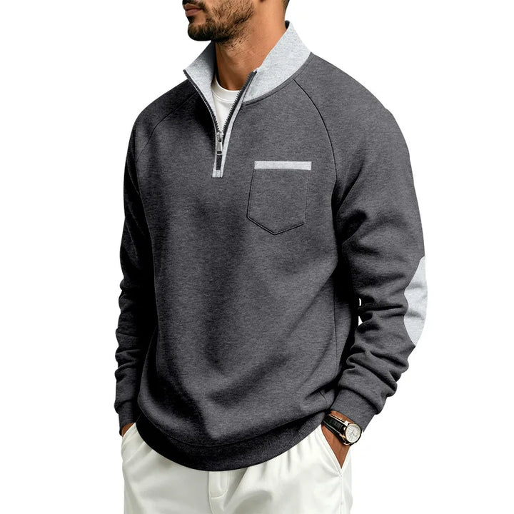 Man wearing a dark gray quarter-zip pullover with a light gray collar, a chest pocket, and elbow patches, posing with hands in pockets.