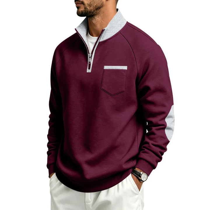 Man wearing a burgundy quarter-zip sweatshirt with a gray collar, chest pocket, and elbow patches, posing with hands in pockets.