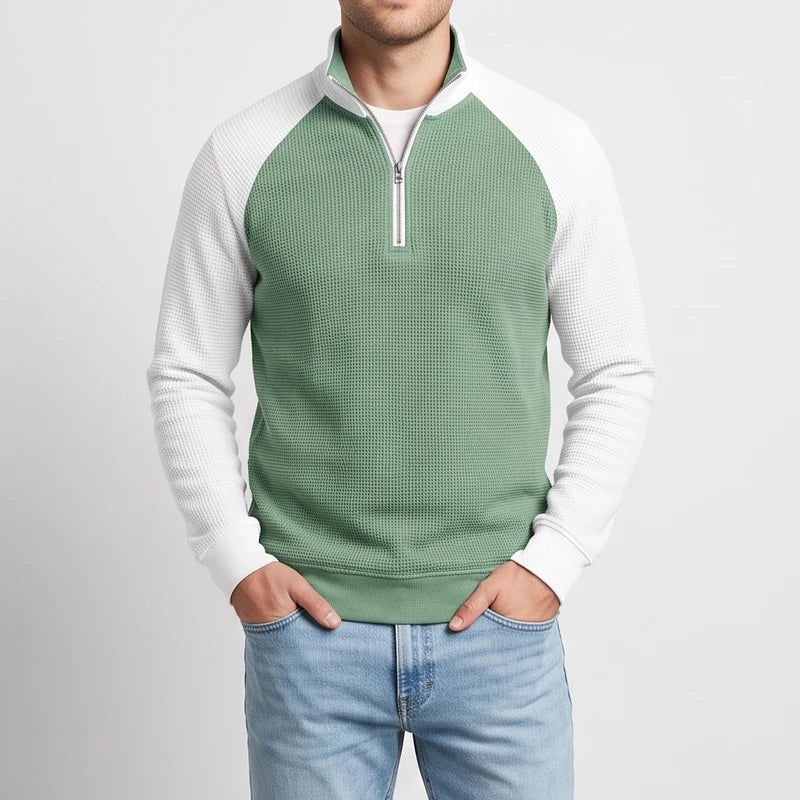Man wearing a two-tone quarter-zip sweatshirt with a sage green waffle knit body and white textured sleeves, hands in pockets.