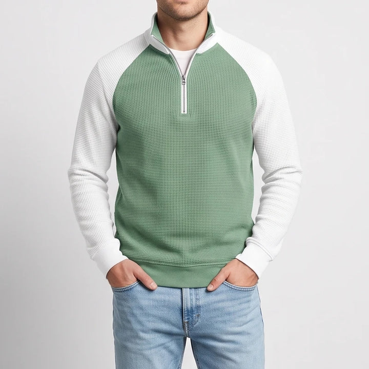Man wearing a two-tone quarter-zip sweatshirt with a sage green waffle knit body and white textured sleeves, hands in pockets.
