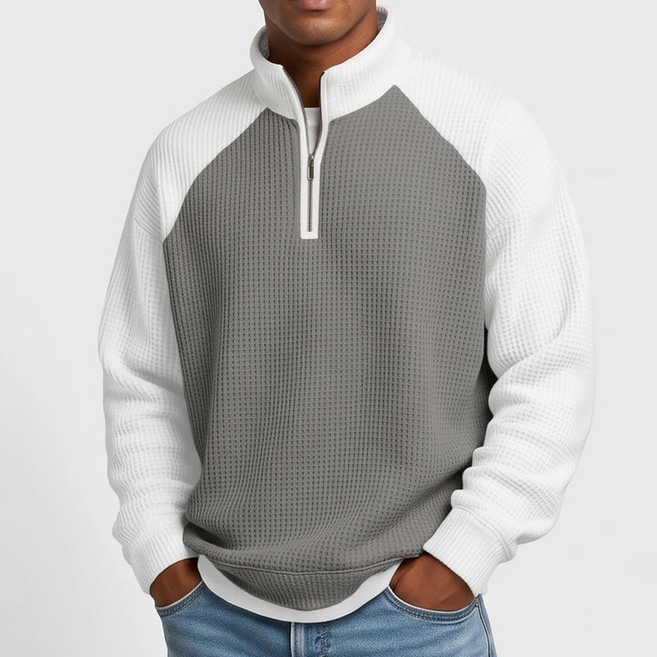 Man wearing a two-tone gray and white waffle-knit quarter-zip sweatshirt with raglan sleeves and light wash jeans.