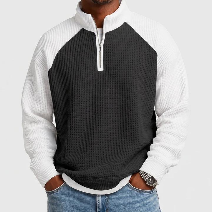 Man wearing a black and white waffle-knit quarter-zip sweatshirt with raglan sleeves and a silver zipper, hands in blue jeans pockets.