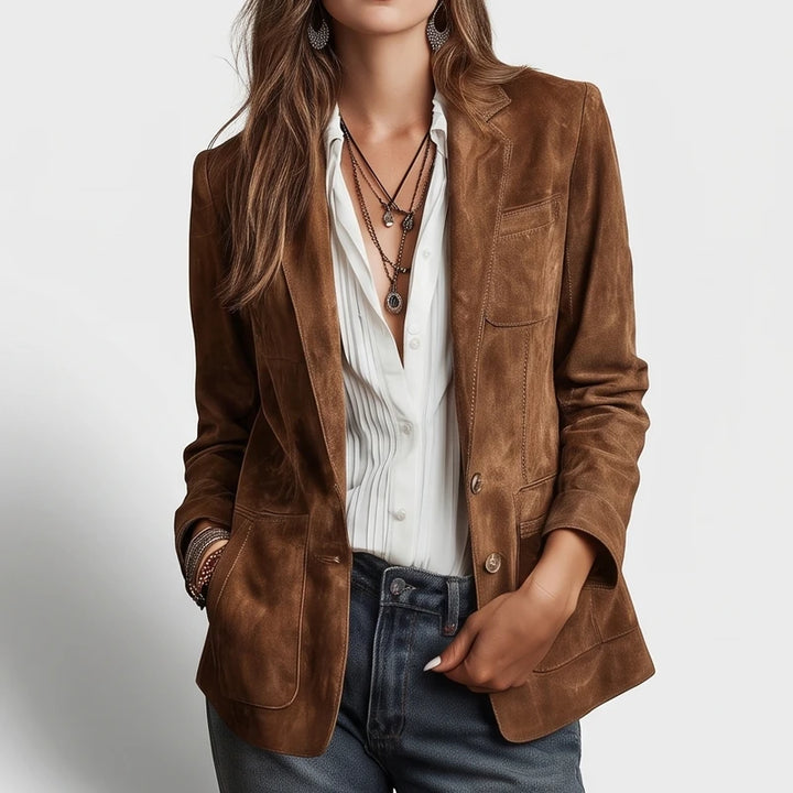 Woman wearing a brown suede blazer with patch pockets, a white pleated shirt, dark blue jeans, and layered necklaces.