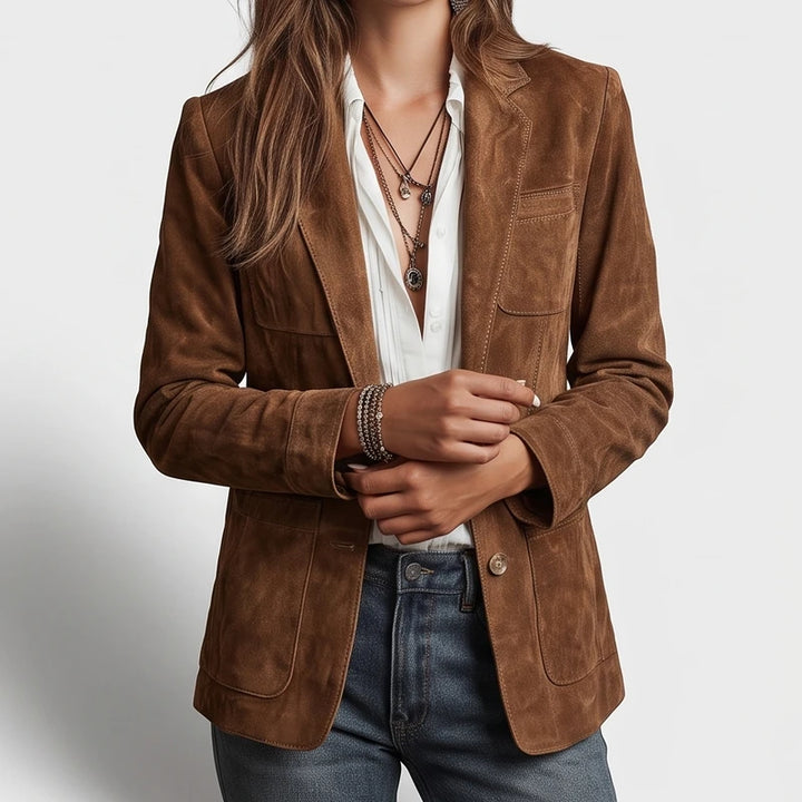 Woman wearing a brown suede blazer over a white shirt, blue jeans, and layered necklaces, hands crossed at the wrist.