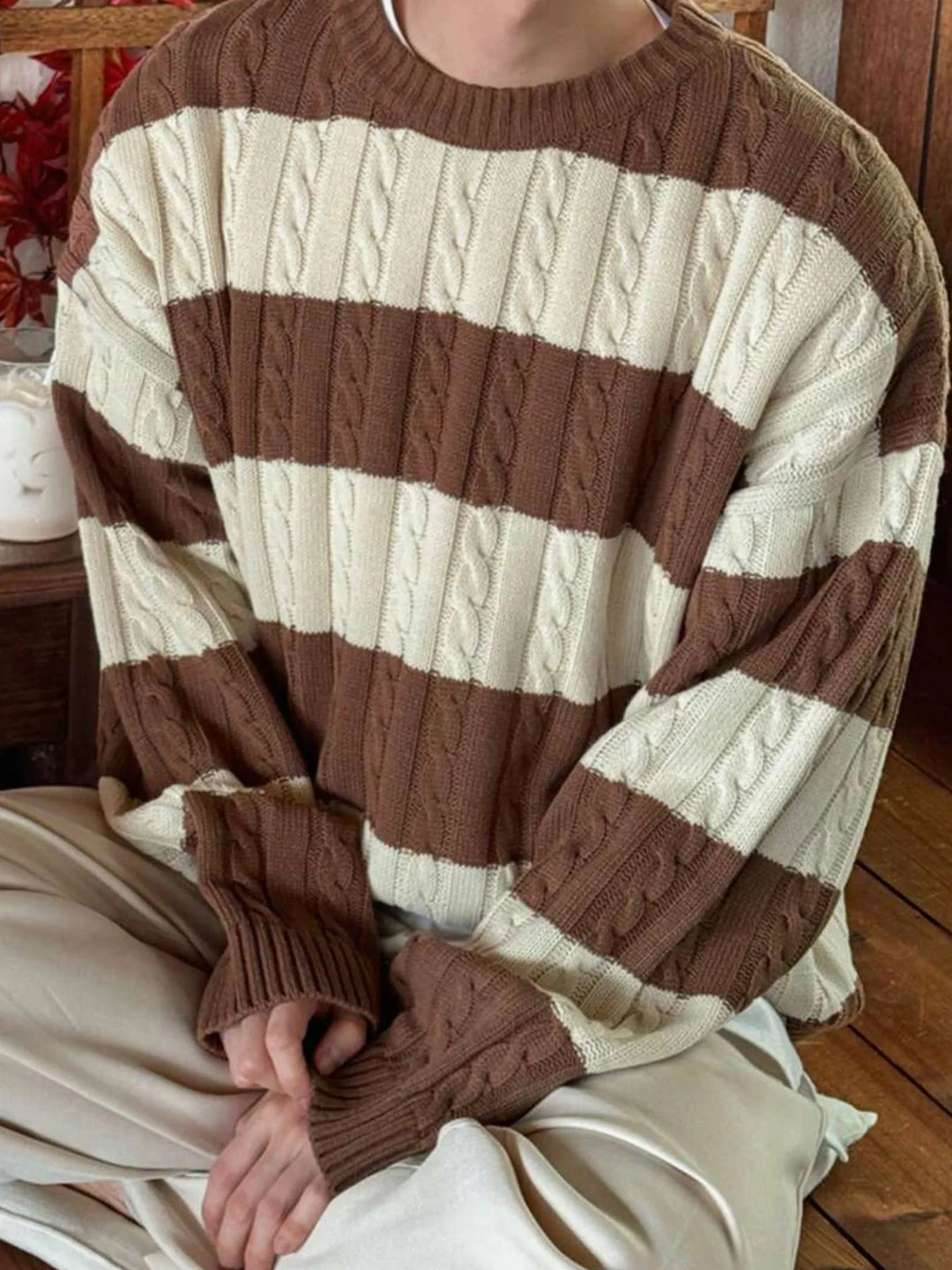 Person wearing an oversized cable knit sweater with broad horizontal cream and brown stripes. Their hands rest on light pants.