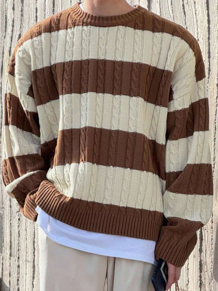 Man wearing a loose-fit cable knit sweater with horizontal brown and cream stripes over a white t-shirt, against a wooden background.