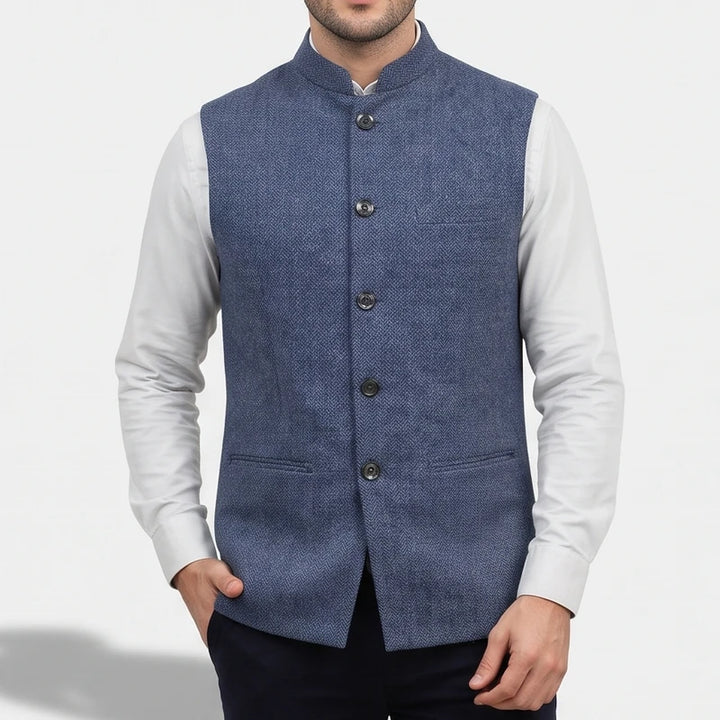 Man wearing a blue herringbone vest featuring a mandarin collar and front buttons over a crisp white shirt.