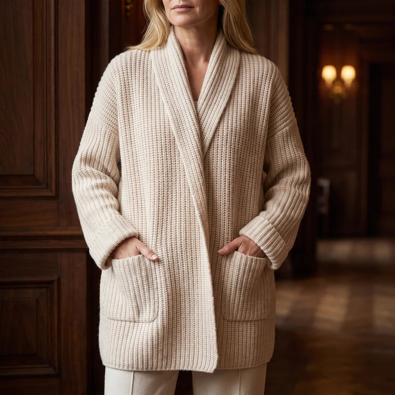 Woman wearing a cream chunky knit cardigan with a shawl collar, ribbed sleeves, and hands in two large front pockets.