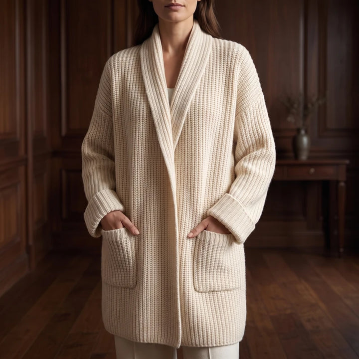 Woman wearing a long, cream chunky knit cardigan with a wide shawl collar, cuffed sleeves, and large front pockets.