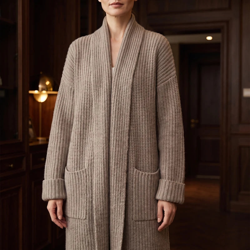 Woman wearing a long, light brown chunky knit cardigan with a shawl collar, two front pockets, and cuffed sleeves.