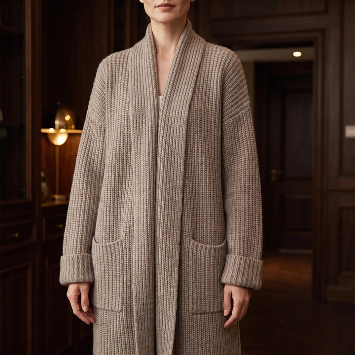 Woman wearing a long, light brown chunky knit cardigan with a shawl collar, two front pockets, and cuffed sleeves.
