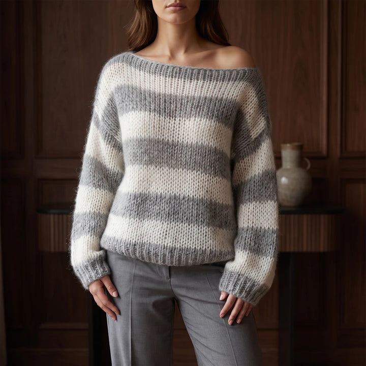 Woman wearing an oversized, off-the-shoulder, striped sweater in gray and cream, paired with gray trousers against a wooden background.