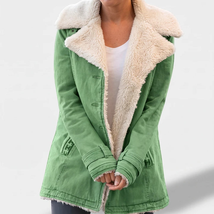 Woman wearing an open green canvas jacket with a fluffy cream sherpa lining and wide lapels, hands clasped.