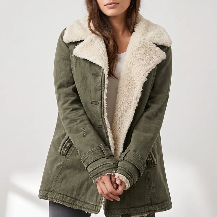 Woman wearing an olive green distressed jacket with a cream sherpa collar and lining, hands clasped, in a neutral setting.