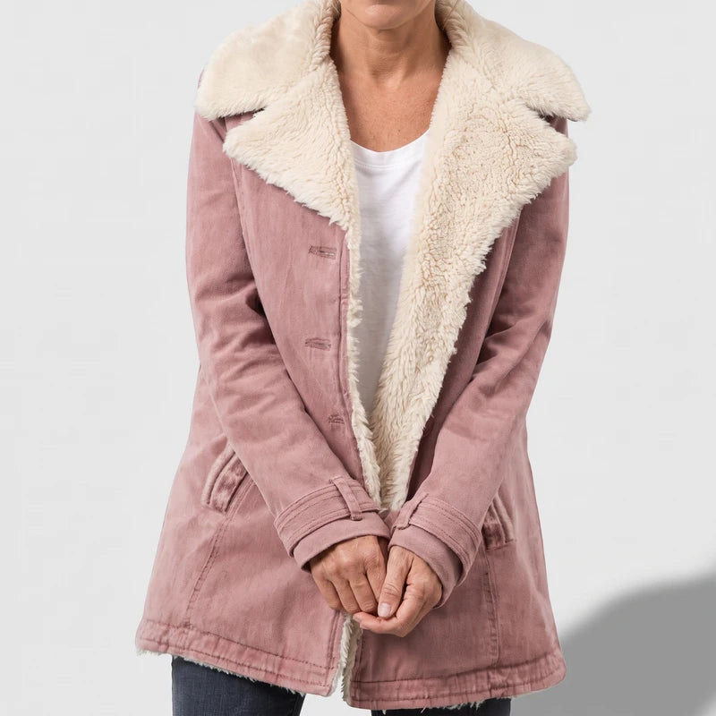 Woman wearing a faded pink sherpa-lined jacket, open to reveal a white t-shirt, standing with hands clasped.