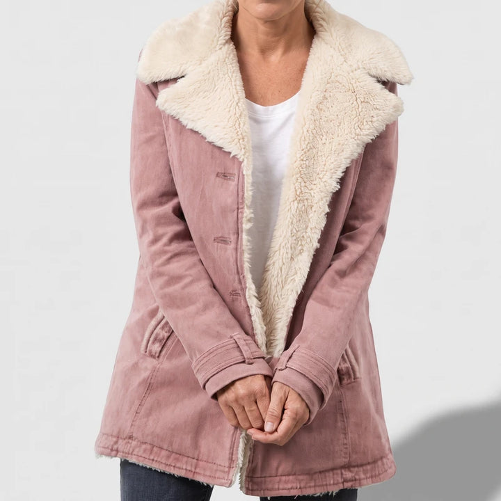 Woman wearing a faded pink sherpa-lined jacket, open to reveal a white t-shirt, standing with hands clasped.