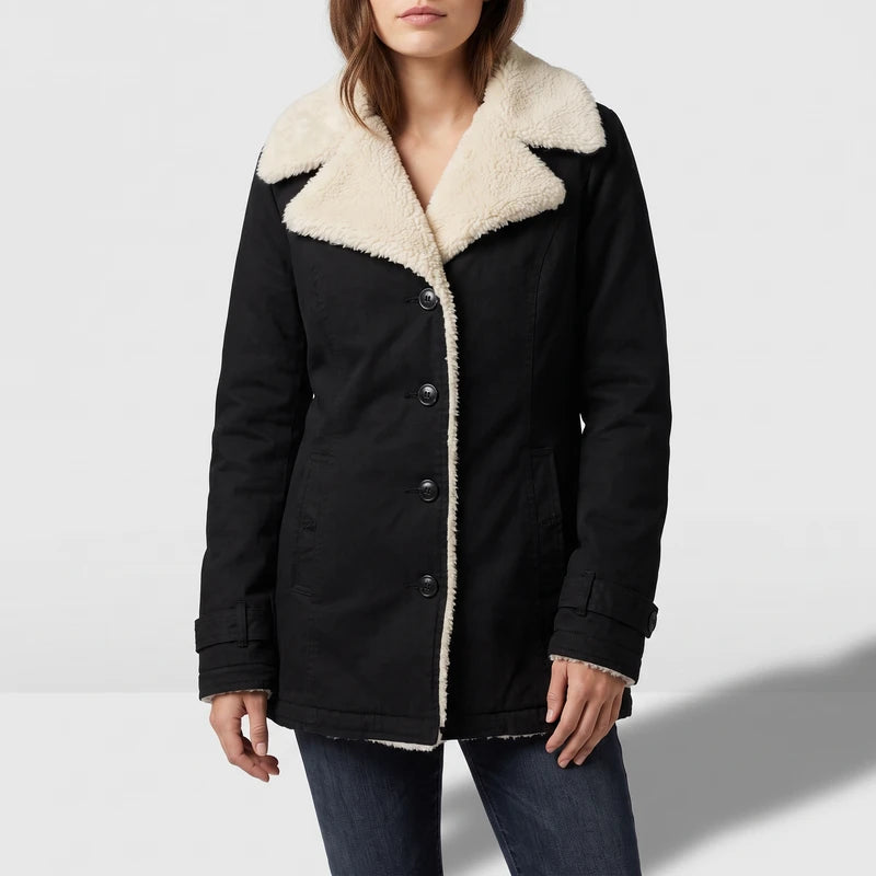 Woman wearing a black coat with cream sherpa lining, black buttons, and cuff straps, over dark blue jeans.