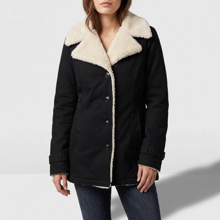 Woman wearing a black coat with cream sherpa lining, black buttons, and cuff straps, over dark blue jeans.
