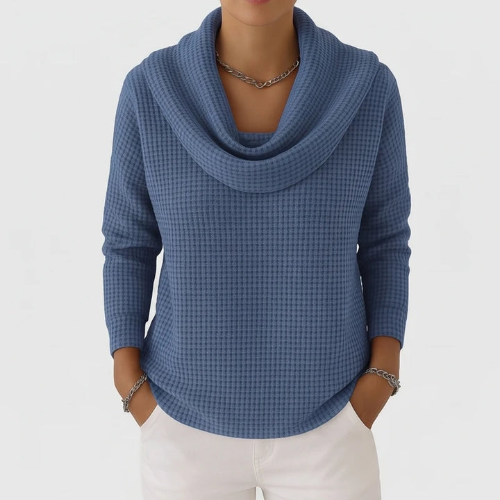 Woman wearing a blue waffle knit sweater with a cowl neck, white pants, and silver chain jewelry.