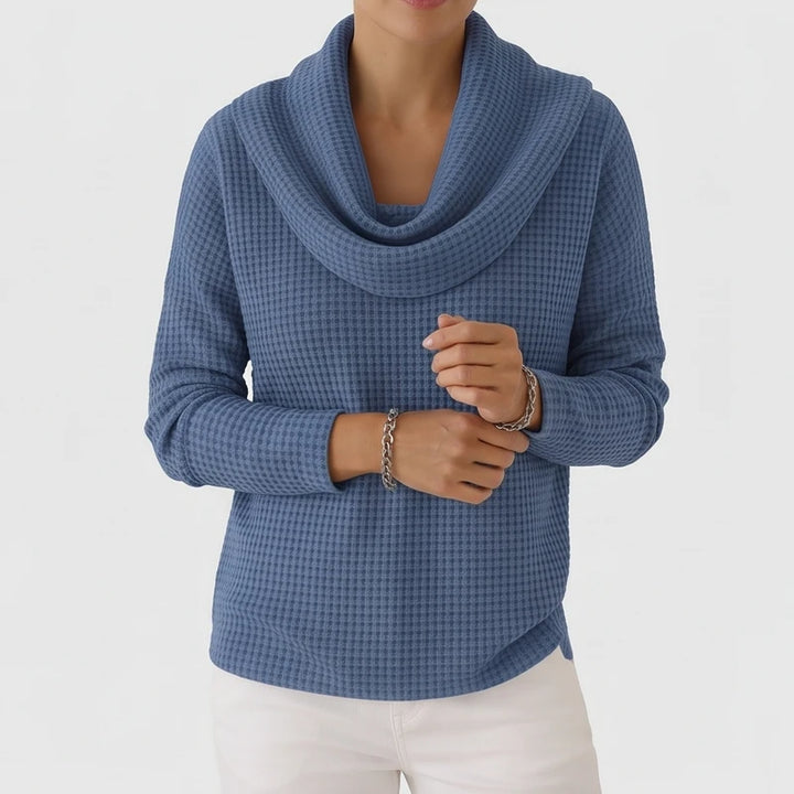 Woman wearing a blue waffle knit cowl neck sweater with a soft drape, light pants, and silver chain bracelets.
