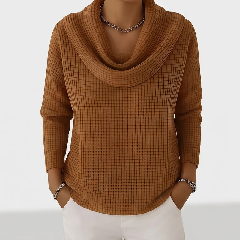 Woman wearing a brown waffle knit cowl neck top with a silver chain necklace and white pants.
