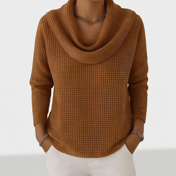 Woman wearing a brown waffle knit cowl neck top with a silver chain necklace and white pants.