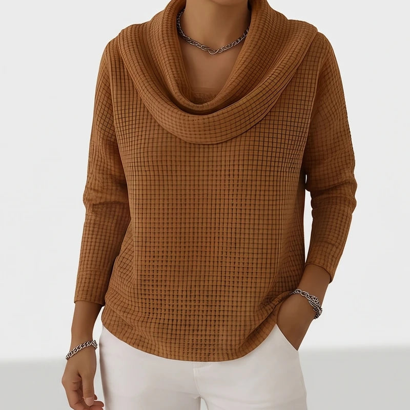 Woman wearing a brown waffle knit cowl neck sweater with a silver chain necklace, posing with hands in white pants pockets.