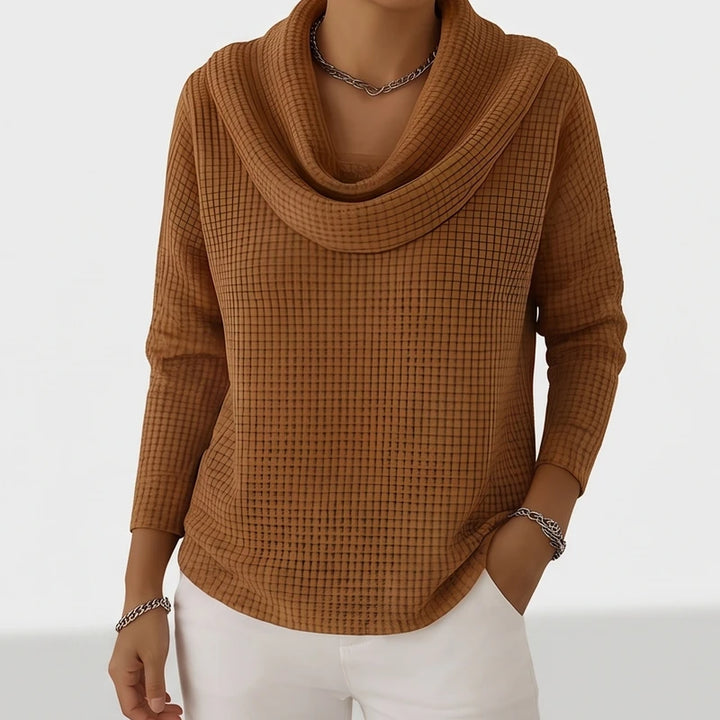 Woman wearing a brown waffle knit cowl neck sweater with a silver chain necklace, posing with hands in white pants pockets.