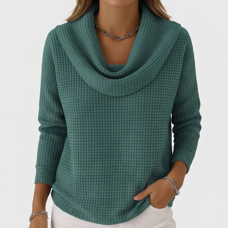 Woman wearing a teal waffle knit cowl neck sweater and white pants, with a silver chain necklace and bracelet.