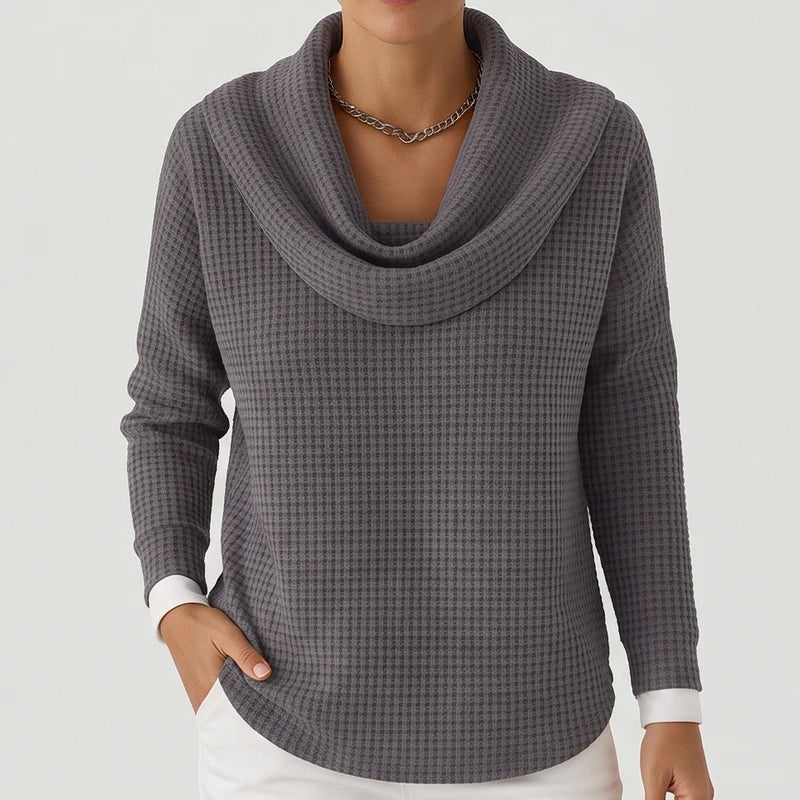 Woman wearing a gray waffle knit sweater with a cowl neck, a silver necklace, and white shirt cuffs peeking out.