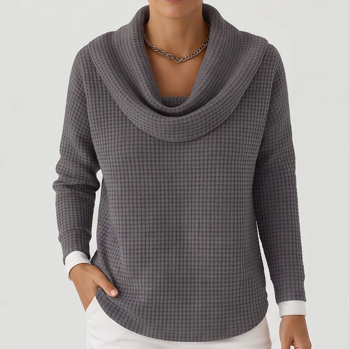 Woman wearing a gray waffle knit sweater with a cowl neck, a silver necklace, and white shirt cuffs peeking out.