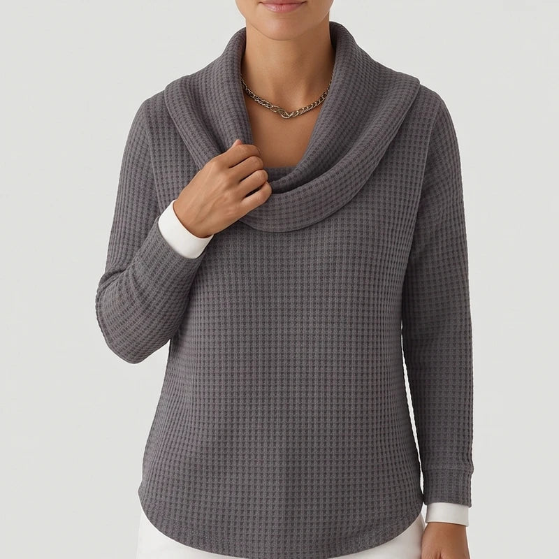 Woman wearing a dark gray waffle knit cowl neck sweater with a curved hem, layered over a white long-sleeve top.